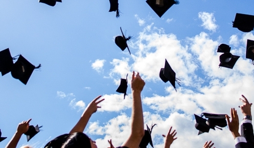 hands tossing up black graduation caps against a blue sky