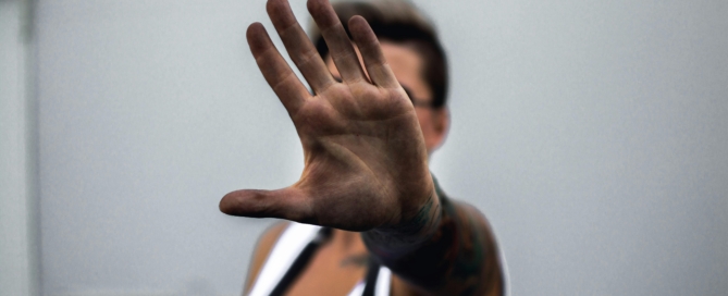 A person wearing a white top holds their hand up toward the camera, palm facing outward, as if signaling stop. The background is neutral and the face is out of focus